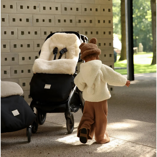 Kind in cremefarbenem Plüschmantel und brauter Mütze schiebt schwarzen Kinderwagen mit weißem Schafsfell-Fußsack Puffmuff