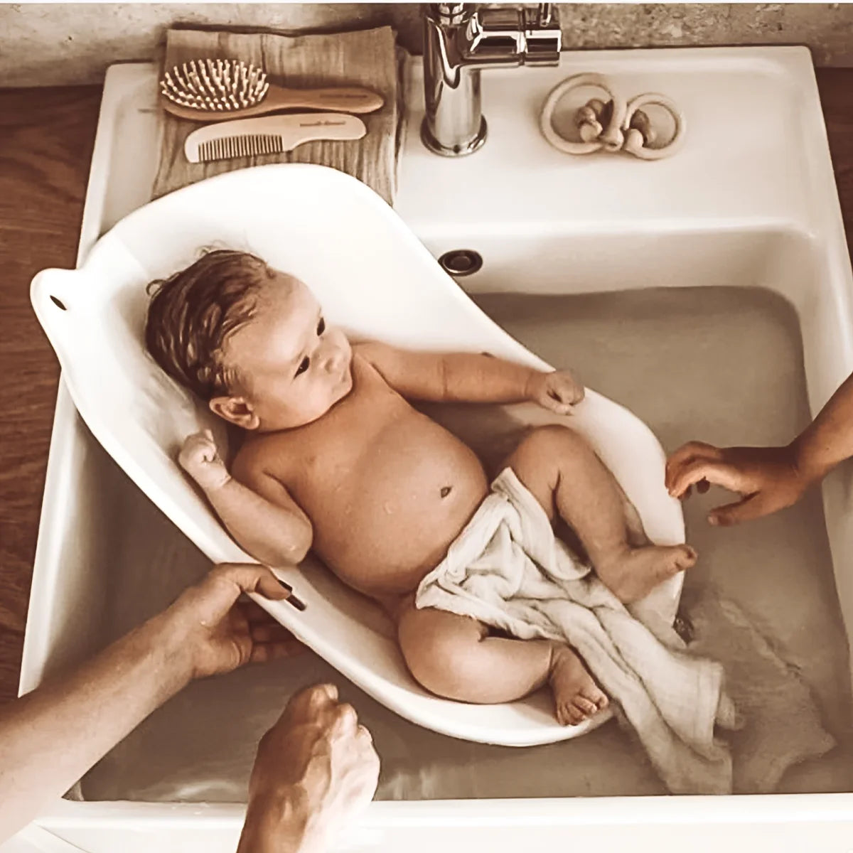 Baby liegt in einem weißen Badeeimer in einer Spüle, Produkt Rassel Wood Naturel