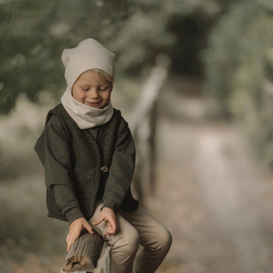 Junge Kind trägt cremfarbenen Beanie und Neck Gaiter, dunkle grüne Weste und hellbraune Hosen