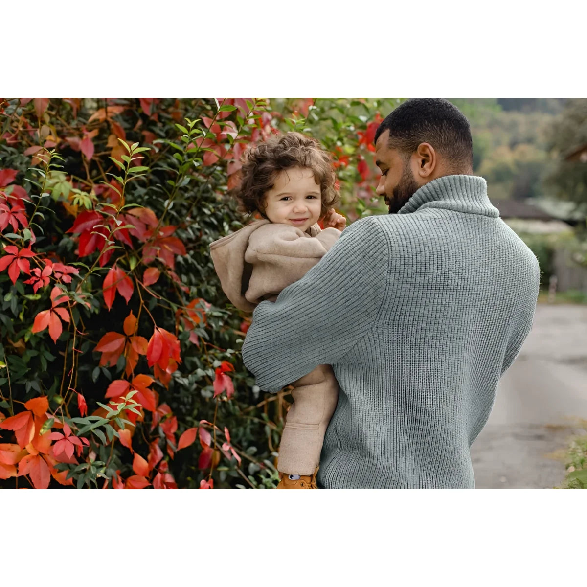 Ein Elternteil in grauem Pullover hält ein Kind neben roten Herbstblättern