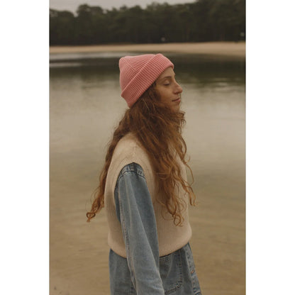 Junge Frau mit langen braunen Wellen, pinker Beanie, helles Sweatervest und Jeans