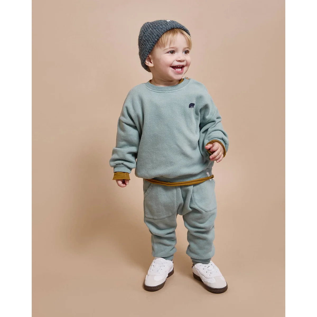 Kleinkind trägt grünem Sweatshirt und Hose, grauer Beanie und weiße Sneaker