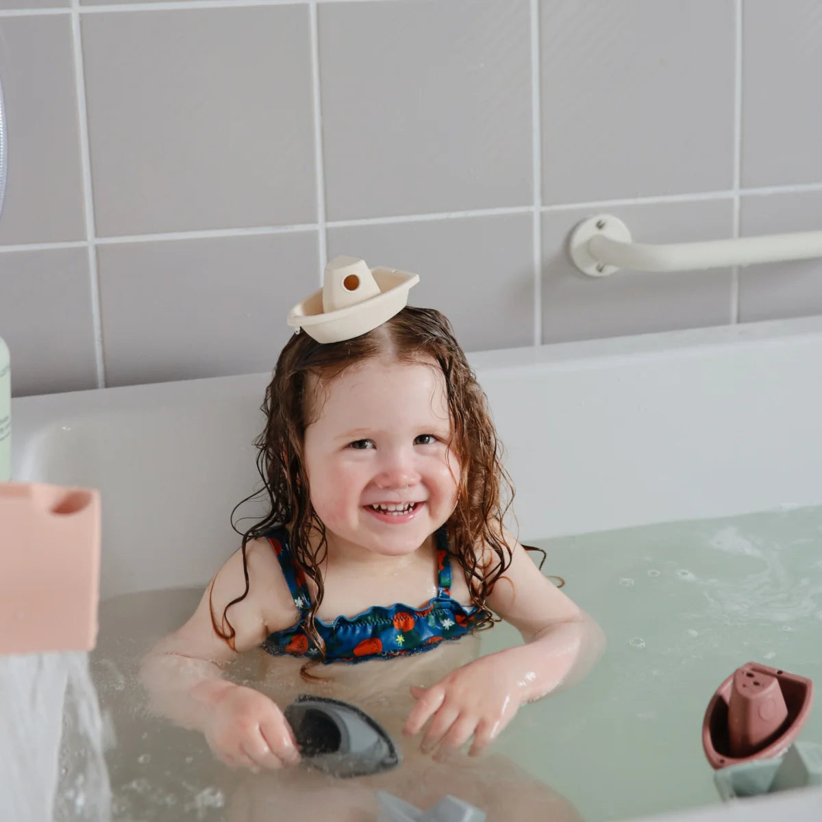 Lächelndes Kind mit lockigem Haar und Cowboyhut in Badewanne mit Badespielzeug