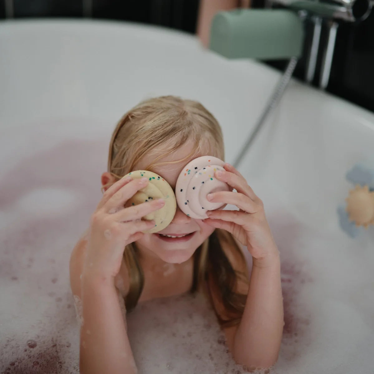 Kind deckt spielerisch die Augen mit bunten Badekugeln im Badewasser