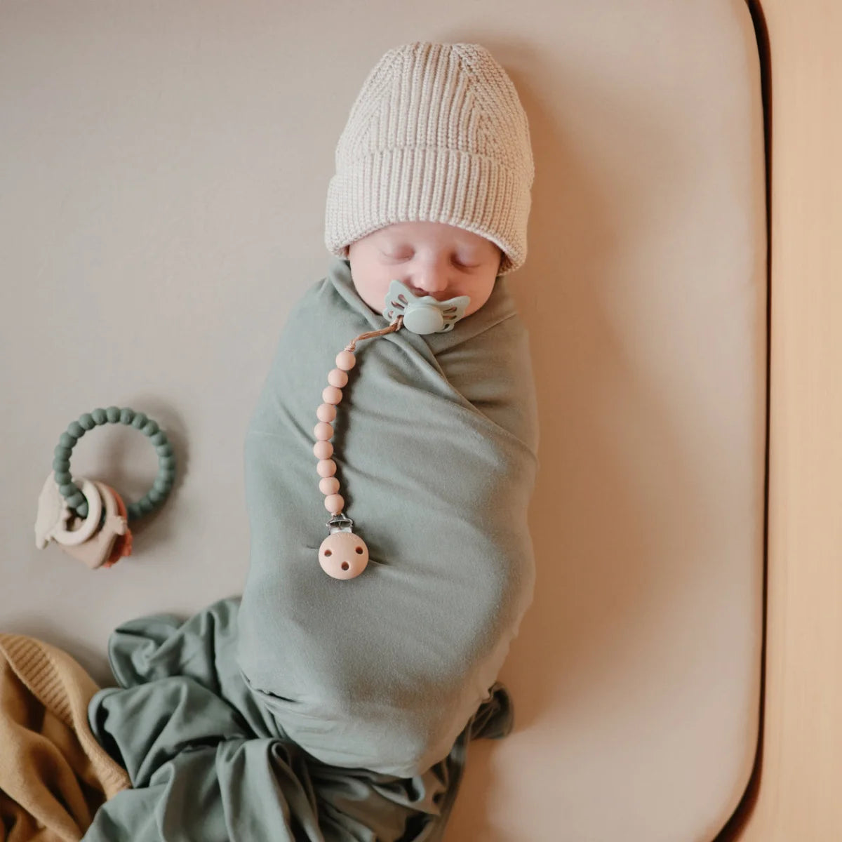 Baby in grüner Swaddle mit Beißring Dino, cremefarbener Mütze und Schnuller