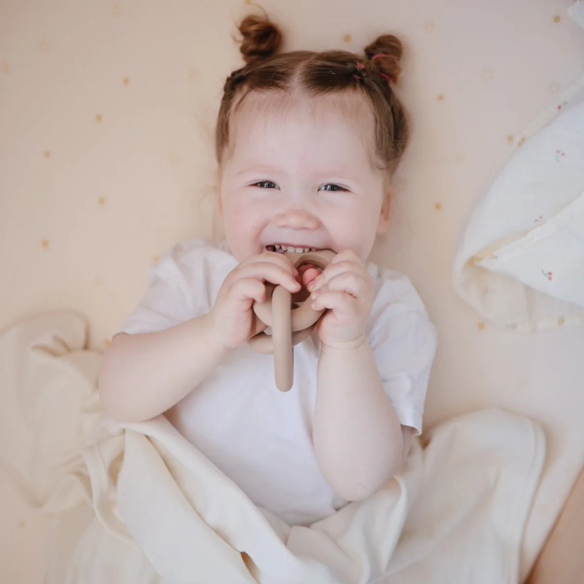 Lächelndes Baby mit zwei kleinen Haarsträußen in weißem Outfit und Beissring Kette