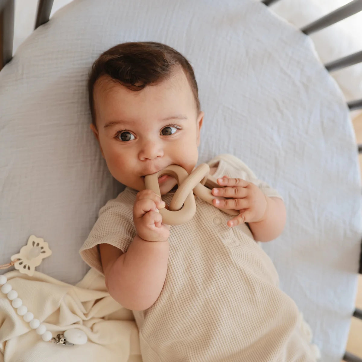 Baby liegt auf einem weißen Kissen und hält ein Holz-Beissring-Spielzeug