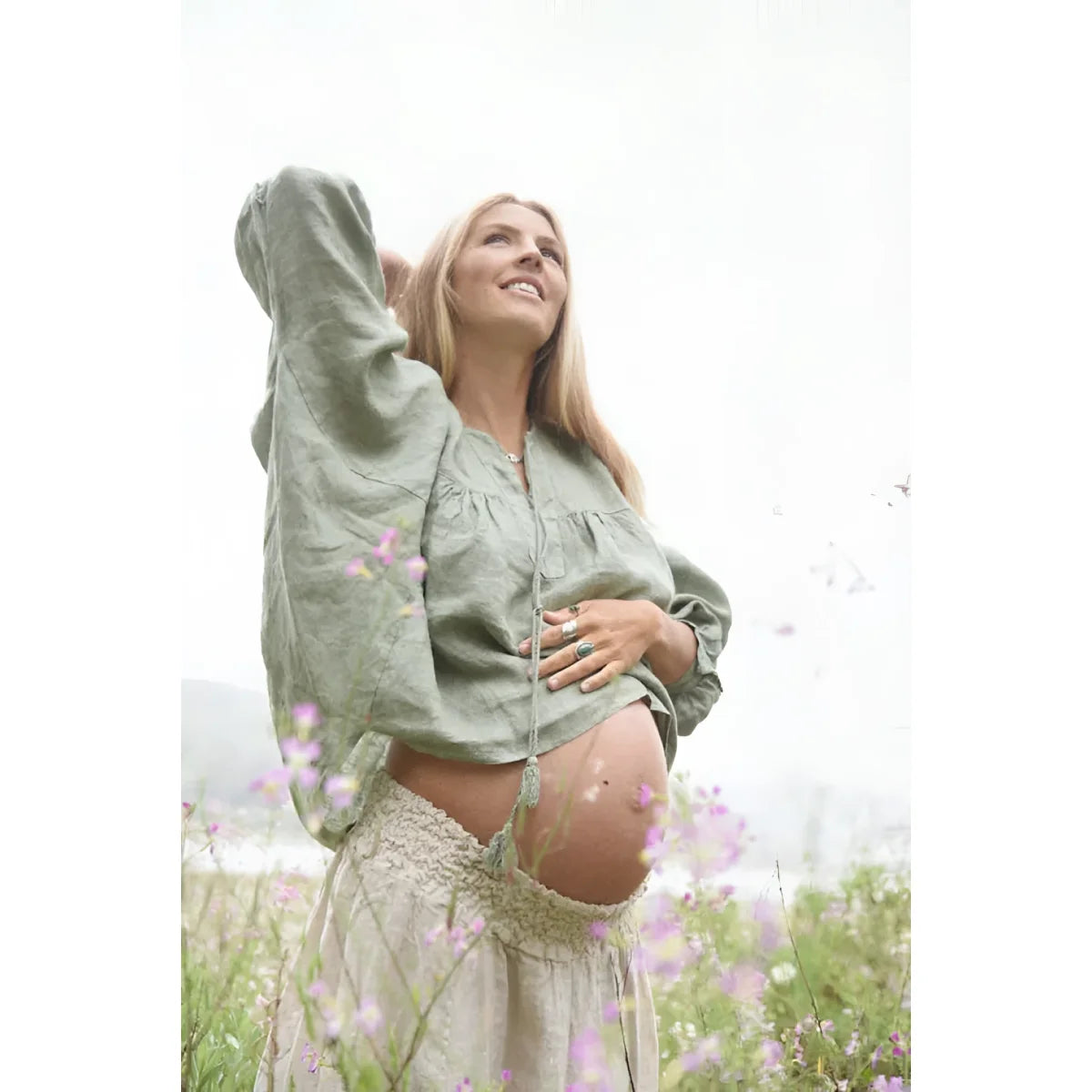 Schwangere Frau in Salbeigrün, umgeben von Wildblumen, präsentiert Bluse Meadow
