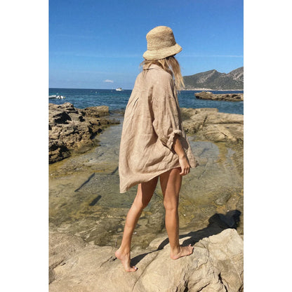 Dame mit beigem Oversized-Hemd und Strohhut auf felsigem Küstengelände