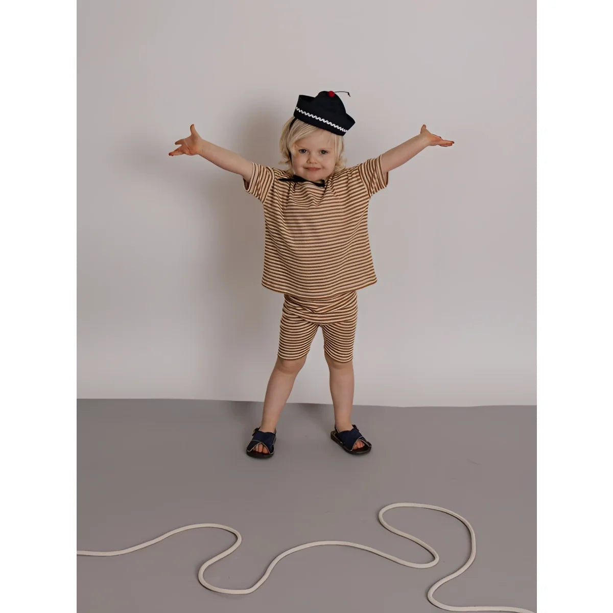 Kind in Boxy T-Shirt Copper Stripes, gestreift beige-braun, marineblaue Sandalen, schwarzer Hut mit weißem Rand