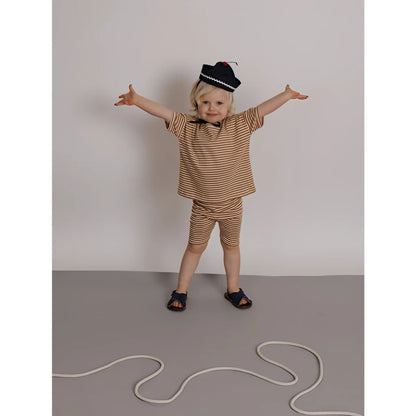 Kind in Boxy T-Shirt Copper Stripes, gestreift beige-braun, marineblaue Sandalen, schwarzer Hut mit weißem Rand