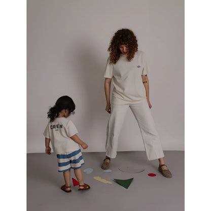 Eine frau trägt ein off white boxy t-shirt mit stickerei und weiten weißen hosen daneben steht ein kind in minimalistischer