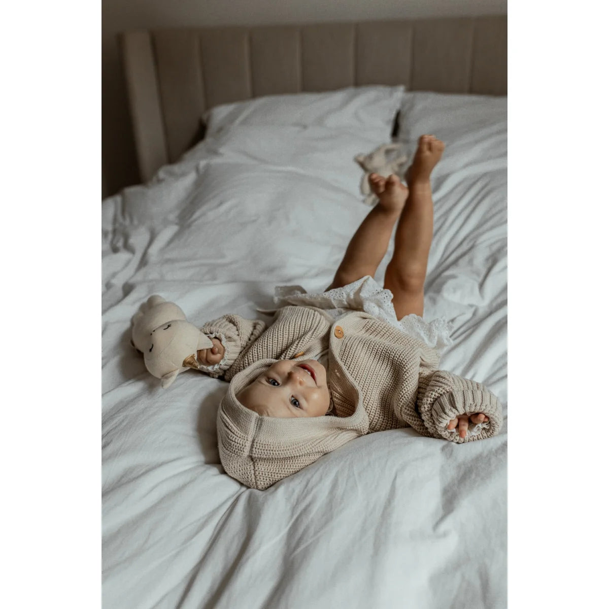 Baby auf einem Bett in einem grob gestrickten Cardigan in Oatfarbe