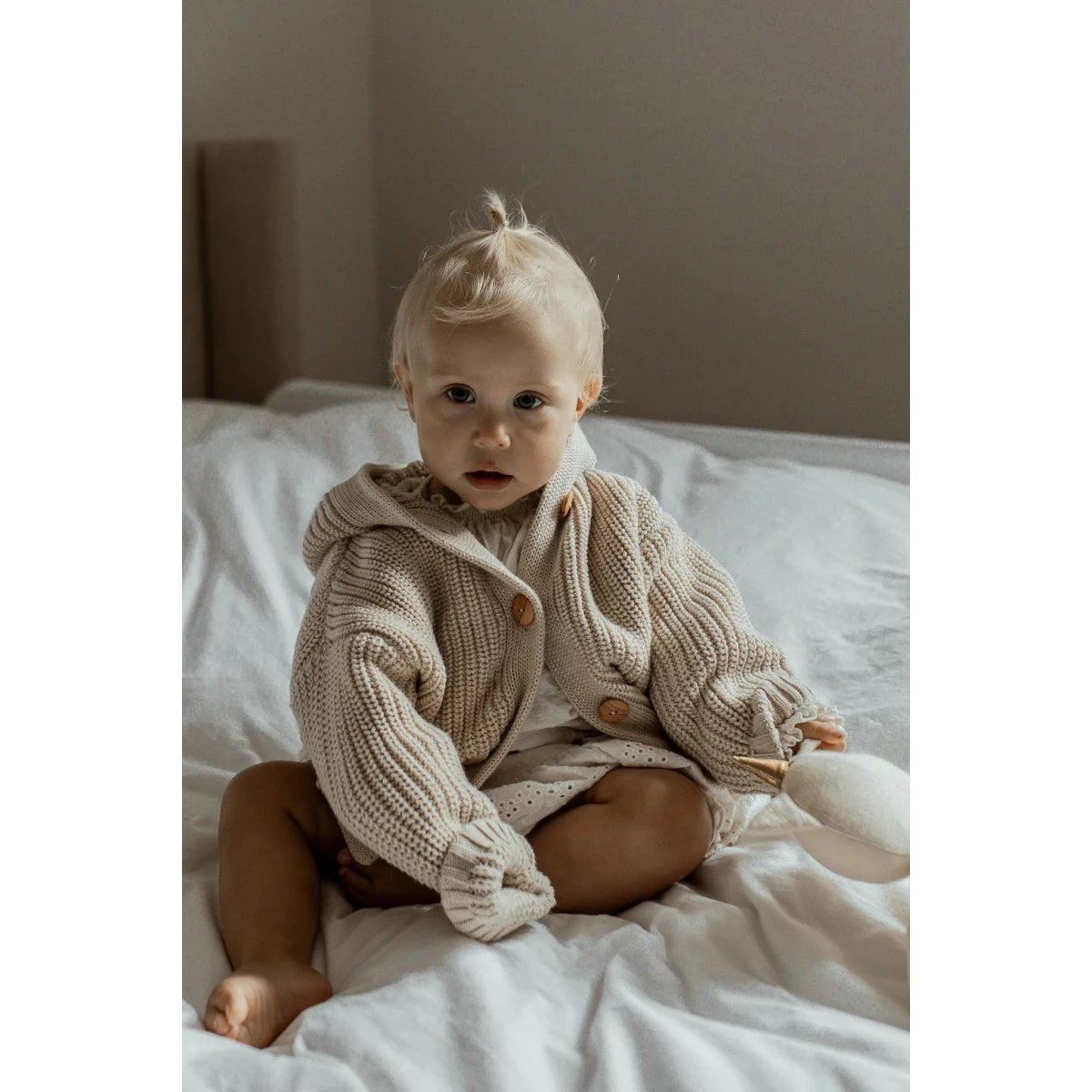 Baby in Chunky Knit Cardigan Oat seated comfortably on a bed