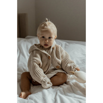 Baby in Chunky Knit Cardigan Oat seated comfortably on a bed