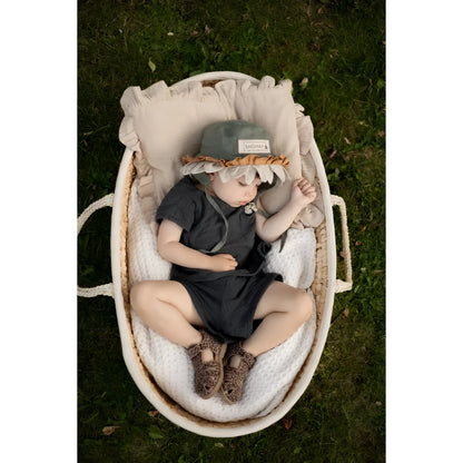 Ein baby trägt einen weichen grauen hut mit gerüschtem hellbraunem saum und sitzt in einem ovalen weidenkorb neben classic