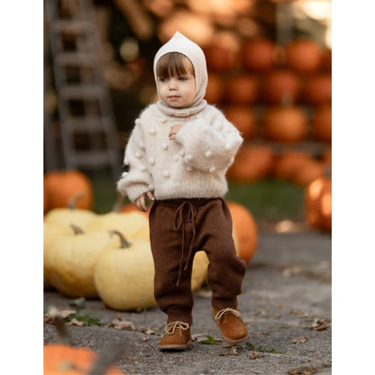 Kleiner Junge in cremefarbenem Pom-Pom-Pullover, braunen Hosen und passenden Schuhen
