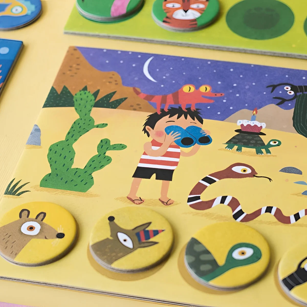 Buntes Kinderrätsel Habitats Lotto: Junge mit Fernglas in Wüste unter Halbmond mit Tieren und Tokens