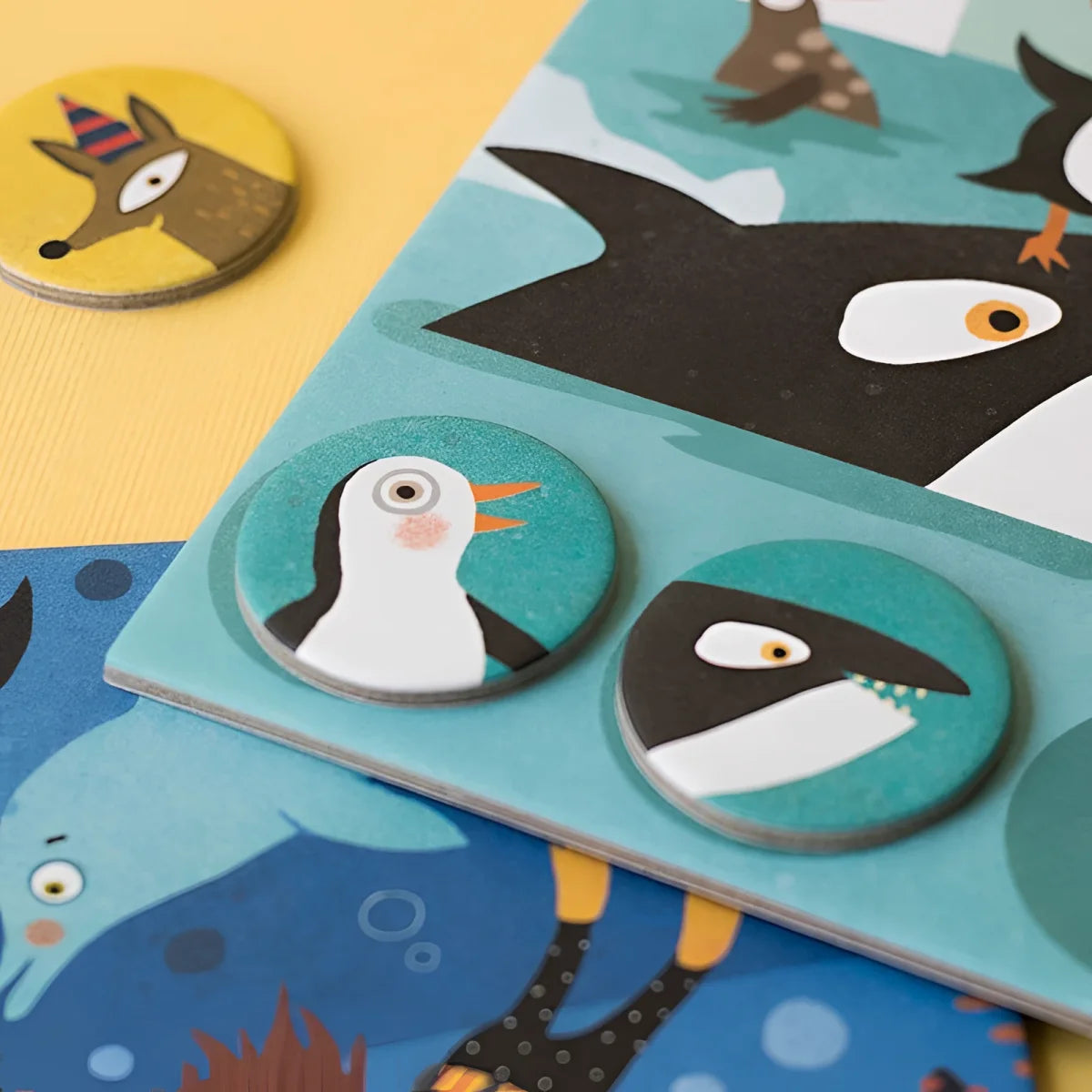 Runde Holzspielmarken mit Pinguin- und Haifisch-Motiven auf türkisem Grund für Habitats Lotto