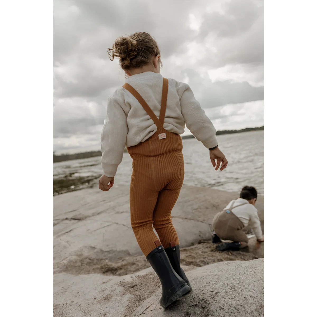 Kind in rostfarbenen Overalls und Gummistiefeln am Strand in Hippy Footless Strumpfhosen
