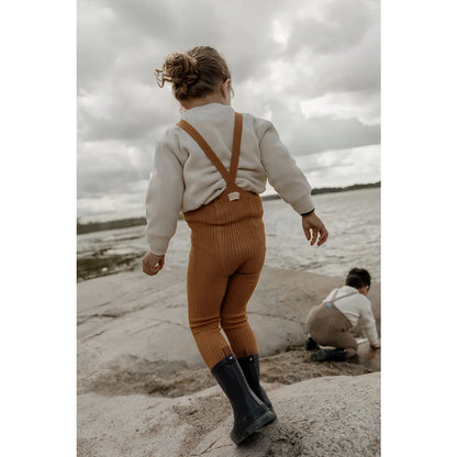 Kind in rostfarbenen Overalls und Gummistiefeln am Strand in Hippy Footless Strumpfhosen