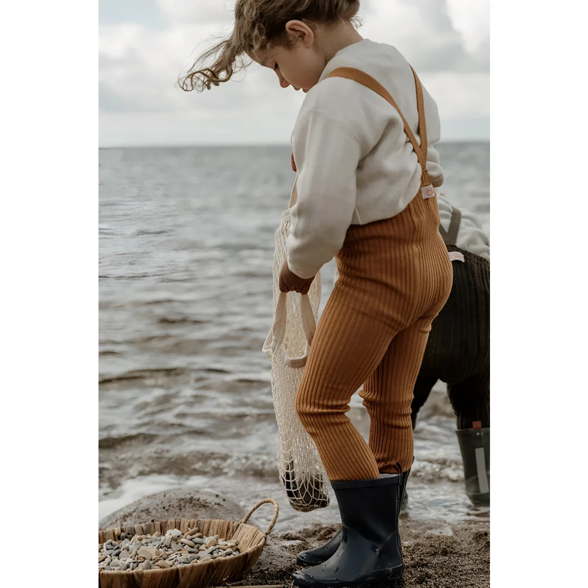 Kind in cremefarbenem Pullover, Cordhosen und Gummistiefeln am Strand