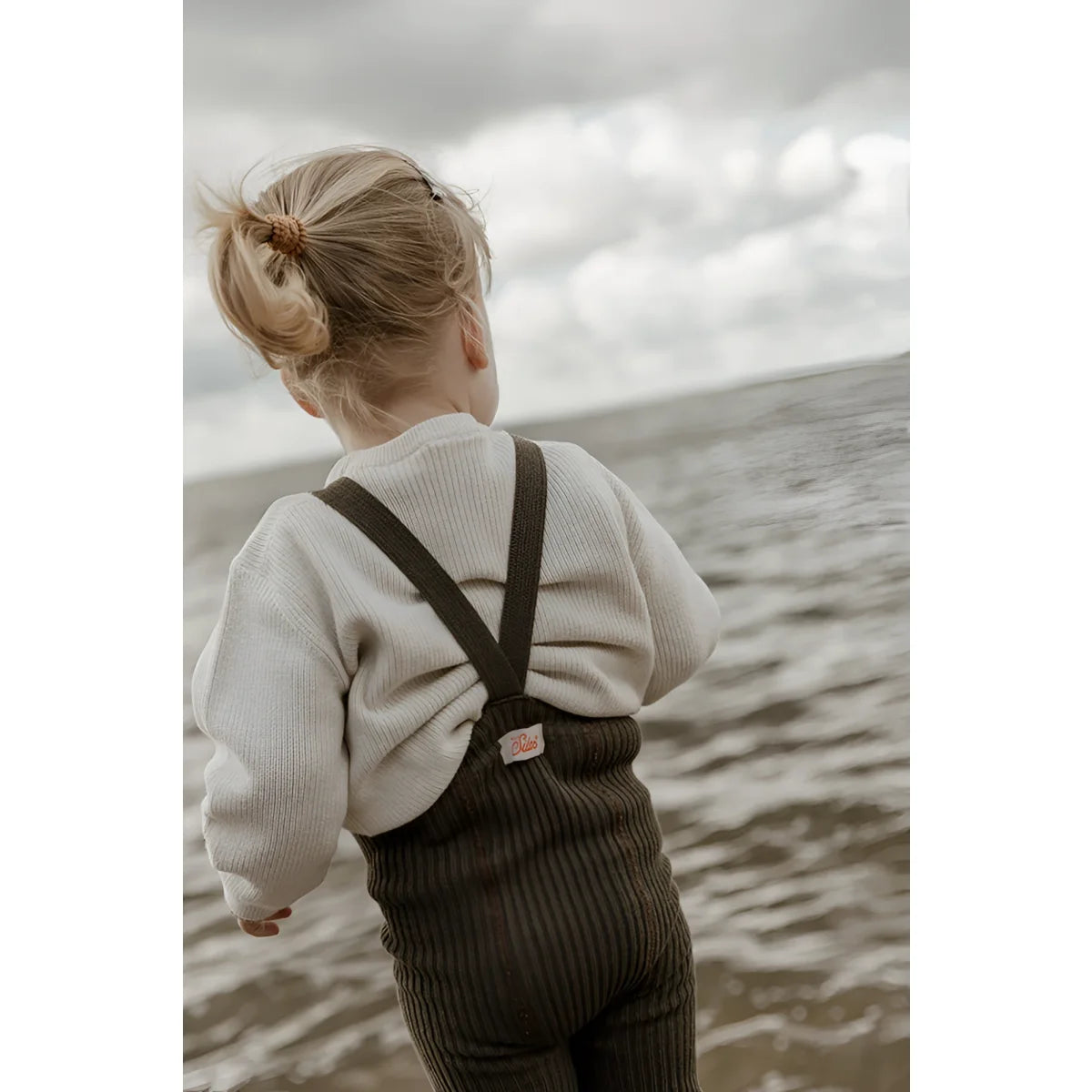 Kind in cremefarbenem Pullover und dunklen Latzhosen am Wasser stehend