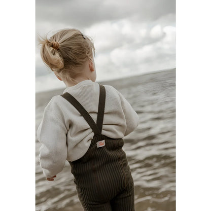 Kind in cremefarbenem Pullover und dunklen Latzhosen am Wasser stehend