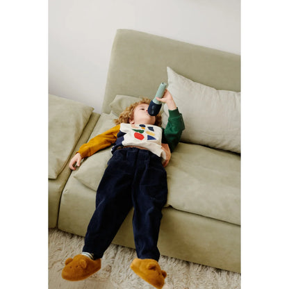Kind tragt navy Corduroy-Hose, weißes Tee mit bunten Formen, senfgelbe Ärmel und Bärenpantoffeln