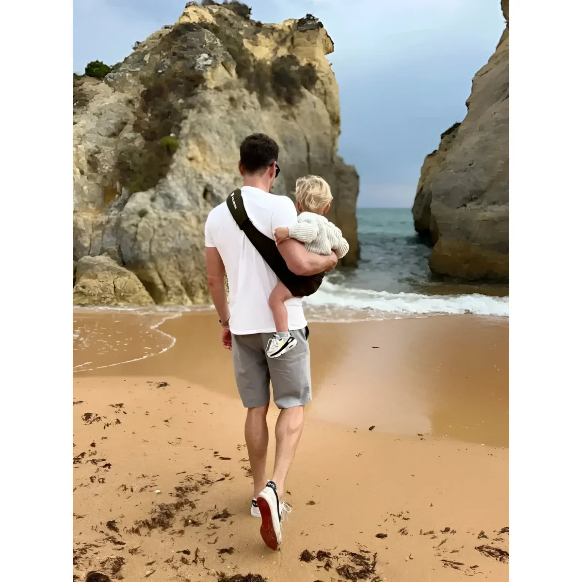 Mann hält Kind am Strand nahe Felsen mit Kindertrage in Armeegrün