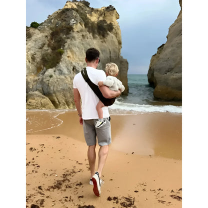 Mann hält Kind am Strand nahe Felsen mit Kindertrage in Armeegrün