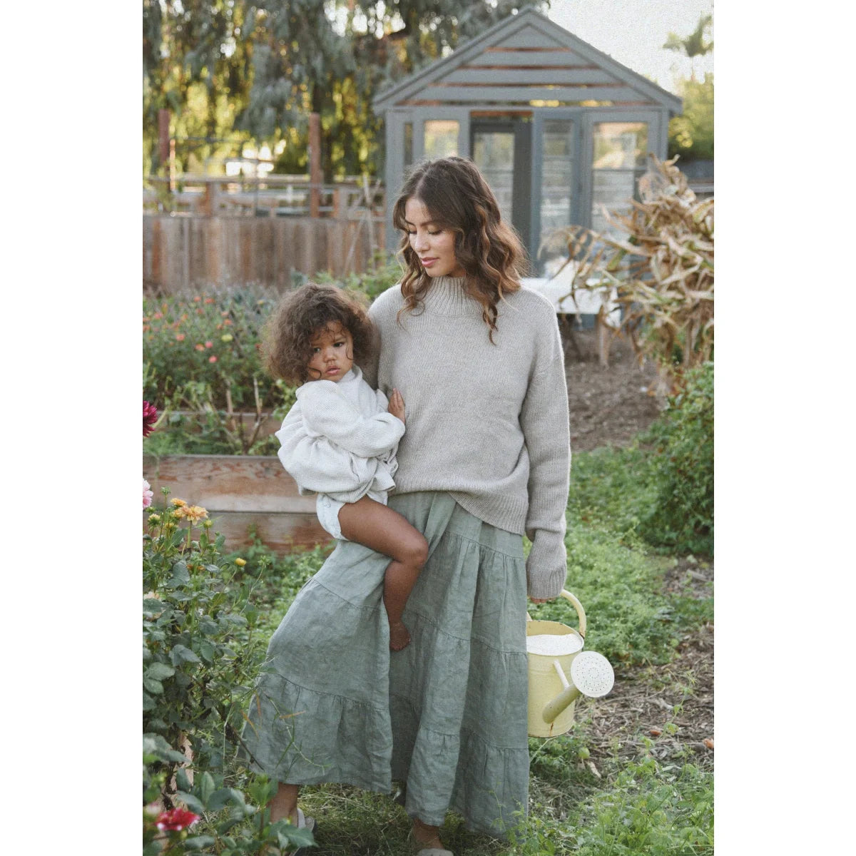 Eine frau trägt das nachhaltige kleid mara eucalyptus in salbeigrün mit einem kind und gießt mit einer gelben kanne