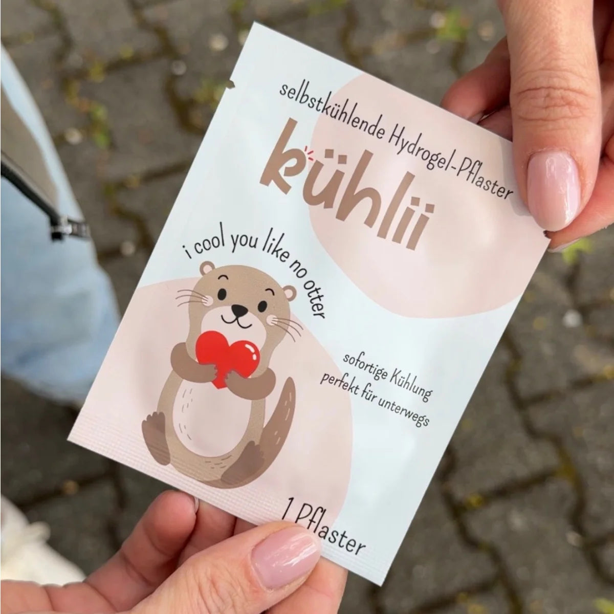 Ein süßer beige brauner cartoon-otter hält ein rotes herz auf einer weißen selbstkühlenden hydrogelpflaster-packung
