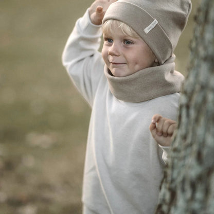 Junger Kind mit beigem Hut und Outfit, schaut hinter Baumstamm hervor