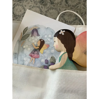 Eine holzfee mit braunen haaren grünem kleid und pfirsichfarbenen flügeln in einer beige tasche aus nachhaltiger babykleidung