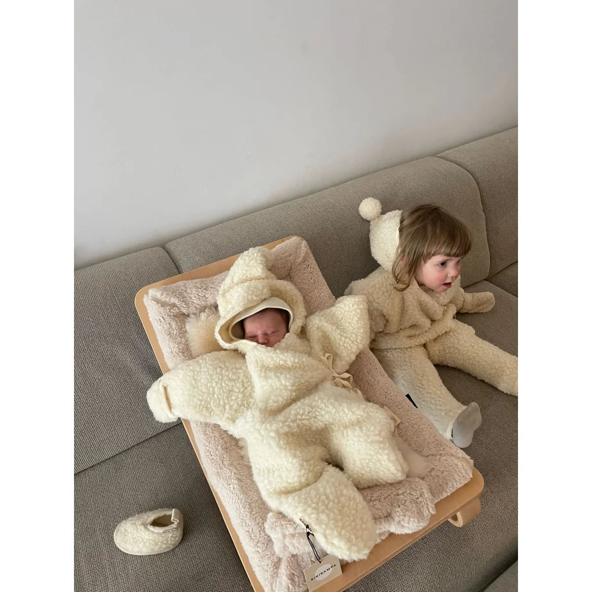 Zwei Kinder in bequemen, cremefarbenen Onesies auf einer Couch im Merino Snugglesuit