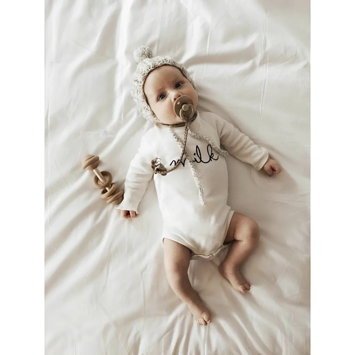 Baby in a white onesie and hat on a white surface with a pacifier, MILK Body Natural