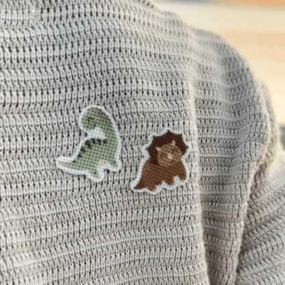 Zwei gestickte Dino-Patches, grün und braun mit weißer Umrandung, auf grauem Strickpullover der nachhaltigen Babykleidung
