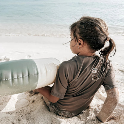 Ein kind trägt einen braunen kurzärmligen rash guard mit ocean child logo und turtle patch am strand mit grün-weißem
