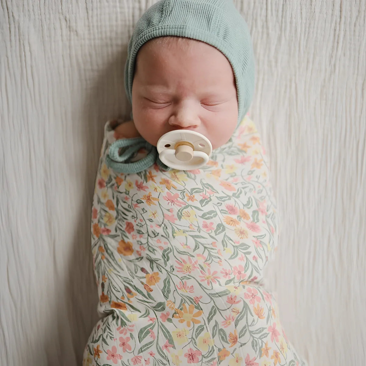Ein neugeborenes baby in einem mulltuch mit pastellblumen in rosa und orange und einer blauen gestrickten mütze