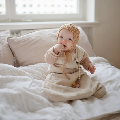 Baby in cream outfit and knitted bonnet on bed with Musselin Schlafsack Fog