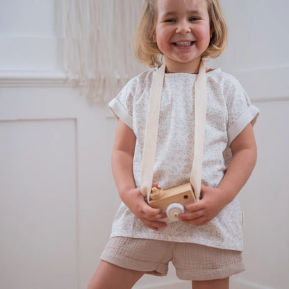 Lächelndes Kind mit Holzspielzeugkamera trägt Musselin Shorts in Beige