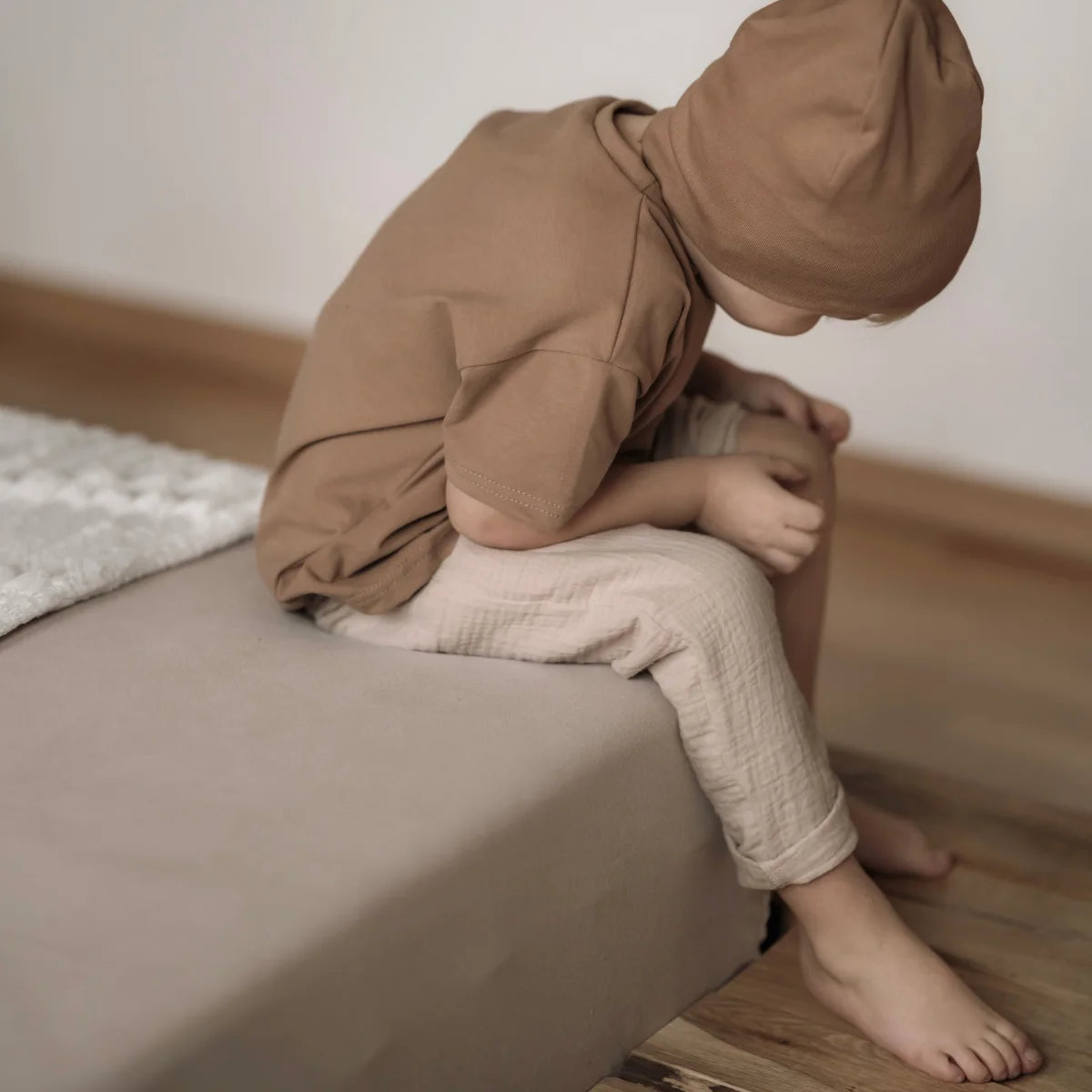 Kind in braunem Kapuzenoutfit, präsentiert die Musselinhose lang in beige