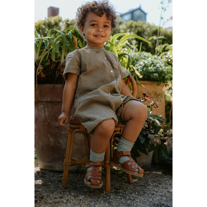 Kind trägt einen weichen olivgrünen leinenstrampler mit textur und knopfleisten zum oscar sandal cognac