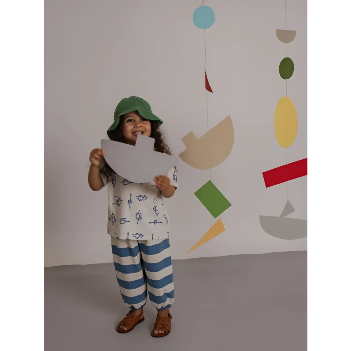 Kind trägt grüne bucket-hut und hält weiße papierschiff-ausschneidung in hosen mit deck-chair-streifenmuster
