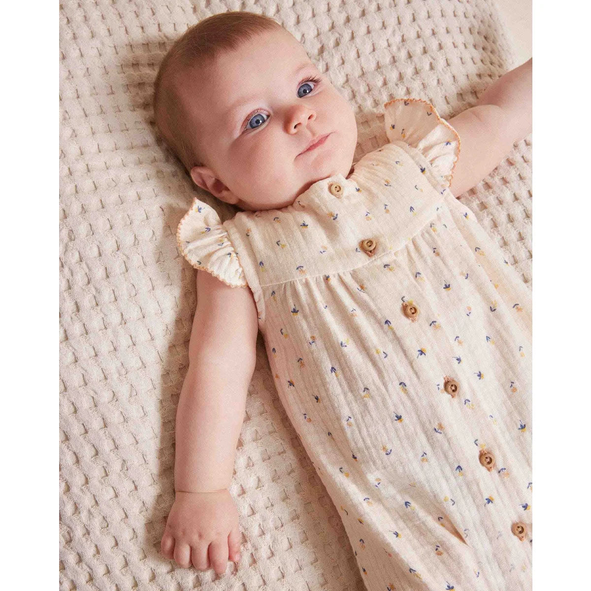 Baby in cream floral jumpsuit with ruffled sleeves and wooden buttons from Unico