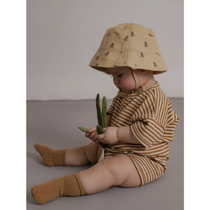 Baby in beigem Bucket Hat mit dunklem Blattmuster trägt Romper Copper Stripes Beach mit passenden Socken