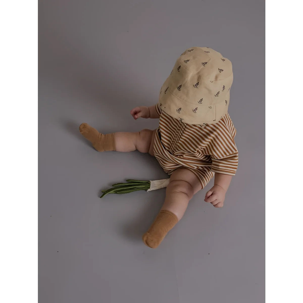 Ein baby trägt einen romper copper stripes beach mit gestreiften braun-beigen shorts und beigem bucket-hut mit blättermuster