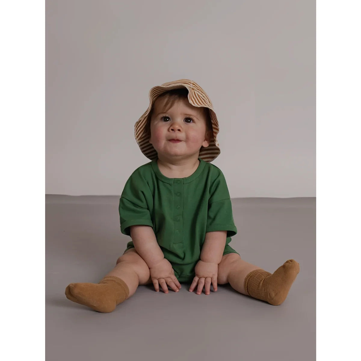 Baby in grüner kurzärmeliger seagrass beach romper aus leichtem stoff mit knopfdetails und gestreiftem strohhut