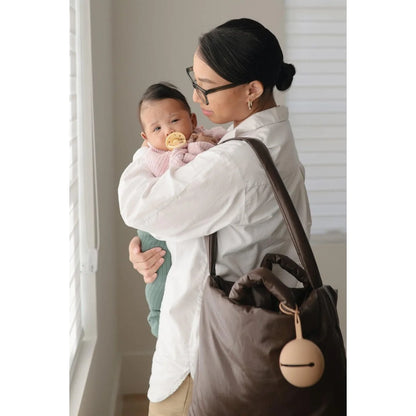 Frau in weißem Mantel hält Baby und Umhängetasche neben Schnuller Etui Shifting Sand